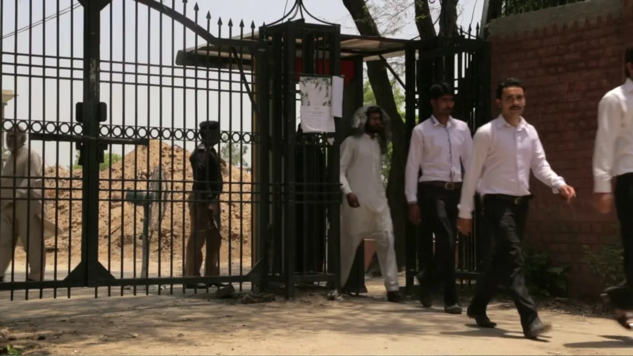 A Girl in the River: The Price of Forgiveness (2015) movie still: fullbody — An outdoor area features a tall black metal security gate beside a red…