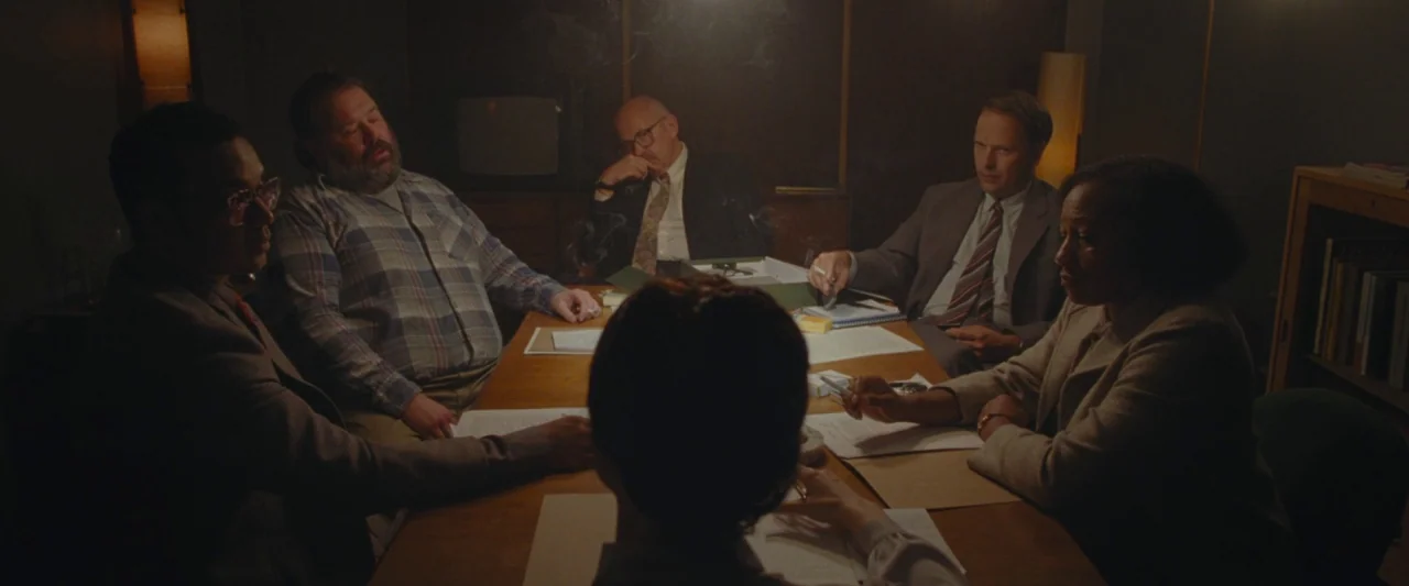 Censor (2021) movie still: wide — A dark, wood-paneled room contains a wooden table with documents and a small television. Five individuals are…