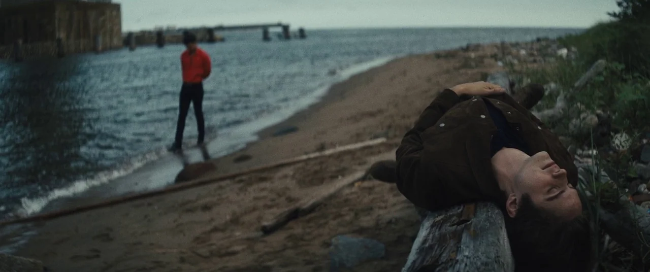 Daniel Isn’t Real movie still: medium — A sandy shore along a body of water with debris and wood scattered nearby. One man lies on a long wooden log…