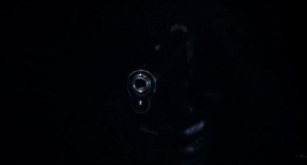 Family Plot (1976) movie still: closeup — A pitch black environment surrounds the front view of a metallic firearm. The muzzle is positioned in the…