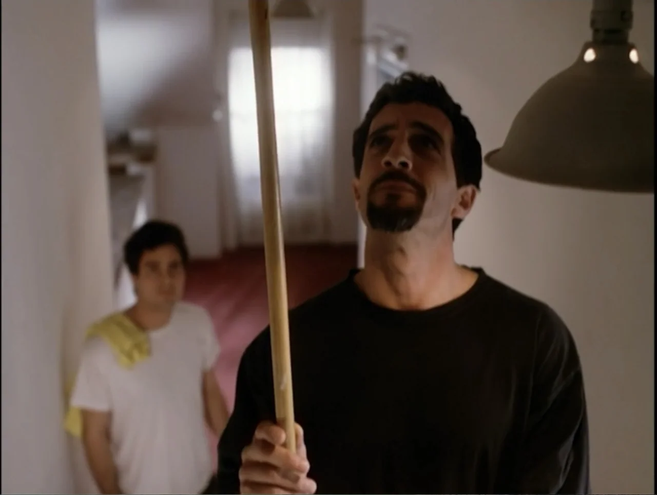 Apartment 12 (2001) movie still: medium — A residential hallway features white walls and a distant window with white curtains. One man stands in the…
