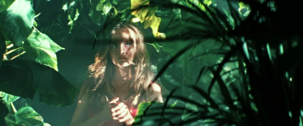 Adoration (2019) movie still: medium — A dense jungle environment with large waxy leaves framing a person in the center. The subject is partially…