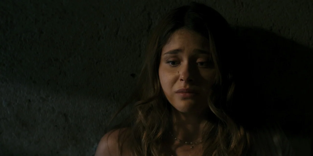My Fault (2023) movie still: closeup — A dark, dimly lit interior space with a textured, shadowy wall surface. A young woman with long hair leans…