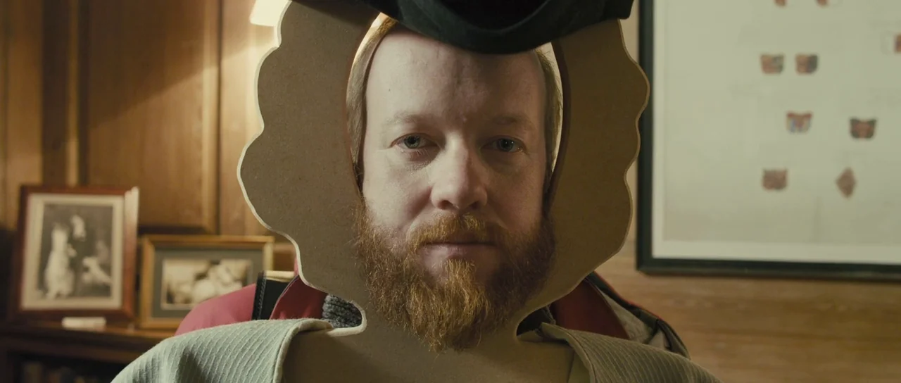 Sightseers (2012) movie still: closeup — A study containing framed pictures on the wall and a wood-paneled wall background. A man holds a cardboard…