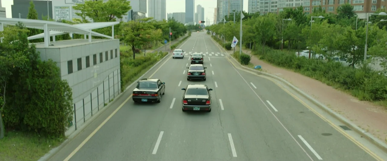 The Gangster, the Cop, the Devil (2019) movie still: wide — An asphalt road stretches forward between lush green trees and modern apartment blocks.…