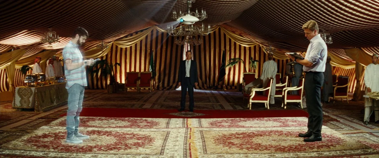 A Hologram for the King movie still: wide — A spacious tented interior features striped fabric walls and ornate rugs on the floor. Three men stand…