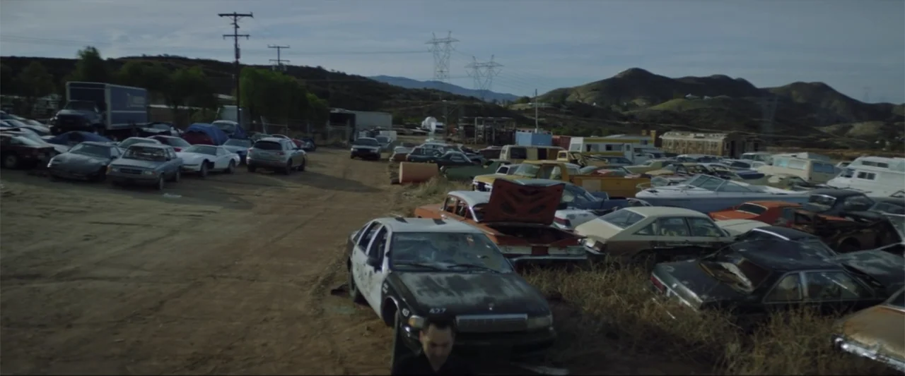 Gender Under Attack (2018) movie still: wide — A sprawling outdoor vehicle graveyard features rows of rusted automobiles and dry grass. Power lines…