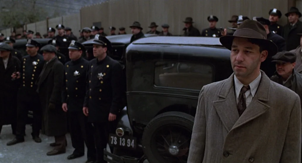 Miller’s Crossing (1990) movie still: medium — A group of men in 1940s formal attire stand near a vintage black automobile. A man in a wool coat and…