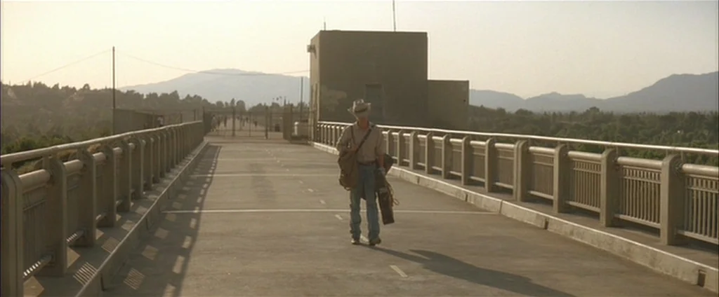 Down In The Valley (2005) movie still: wide — A wide, empty concrete pedestrian bridge overlooks a valley with distant hills at dusk. A lone man…