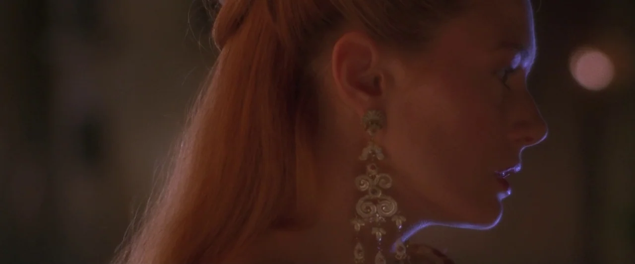 Dangerous Beauty (1998) movie still: closeup — A dimly lit interior space with a dark, indistinct background. A woman in profile wears large…