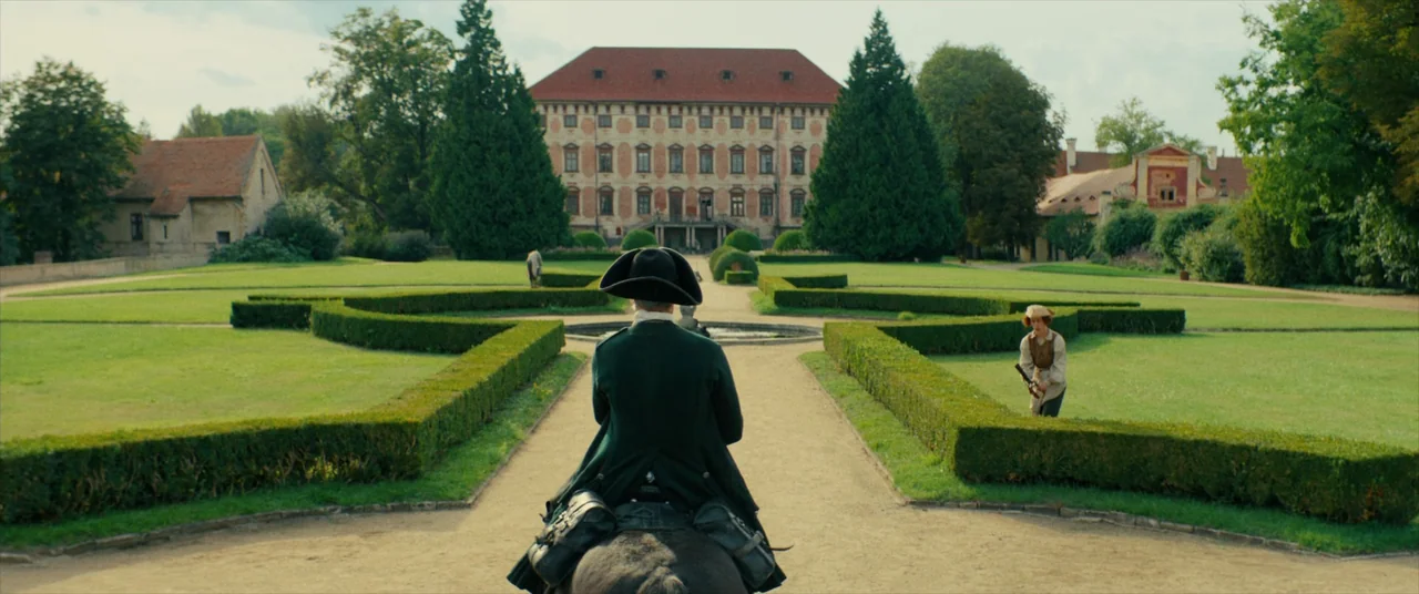 The Promised Land (2023) movie still: fullbody — An expansive estate garden features manicured hedges and a central fountain in front of a manor. A…