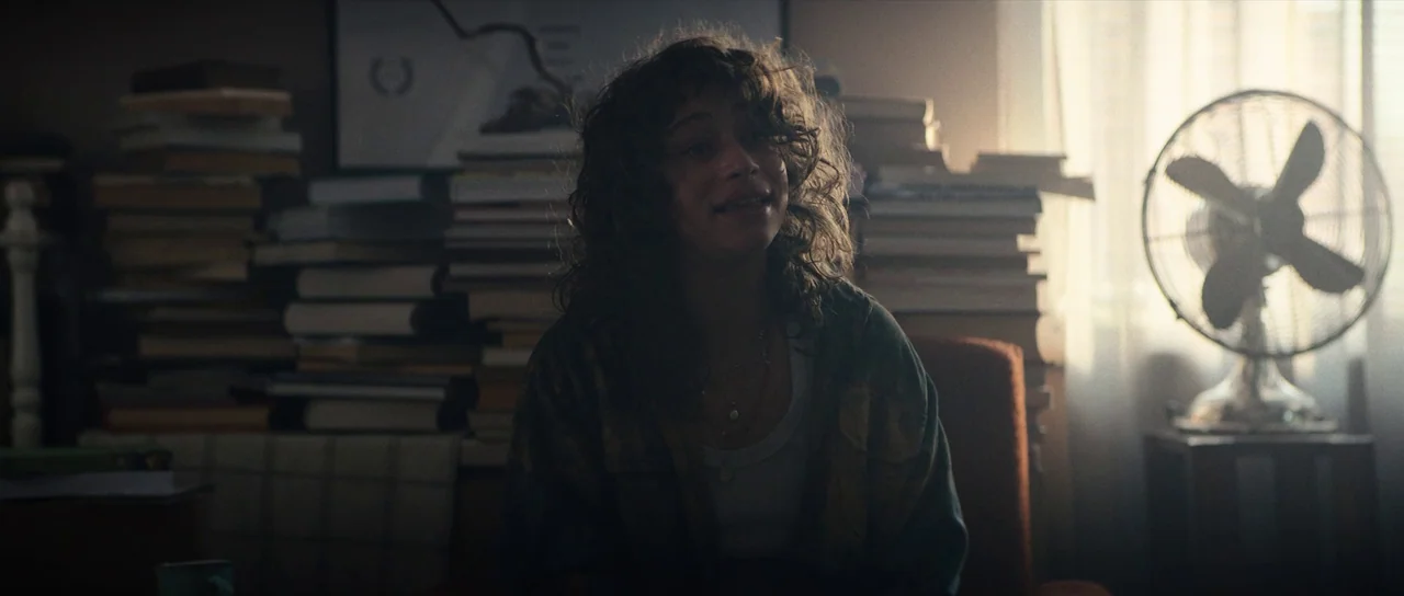 Hellraiser (2022) movie still: medium — A cluttered room features stacks of books lining the wall behind a central figure. A person with curly hair…