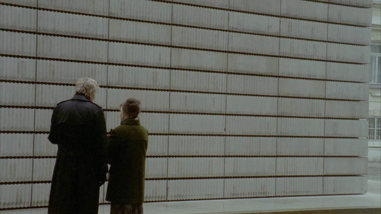 Museum Hours (2012) movie still: wide — A minimalist modern facade made of stacked, horizontal concrete blocks dominates the scene. A man and a…