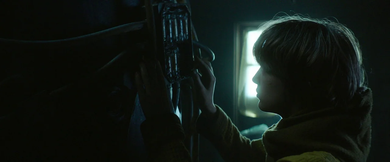 Vesper (2022) movie still: medium — A dimly lit room contains a large dark mechanical apparatus with exposed wires and components. A young woman…