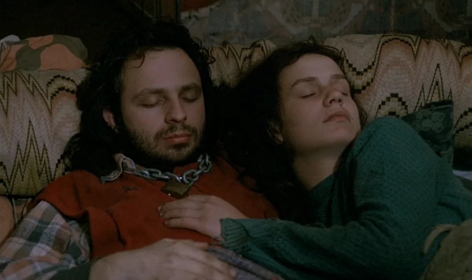 Vagabond (1985) movie still: closeup — A man and woman sleep together on a bed covered in patterned cushions. The man wears a metal chain and…