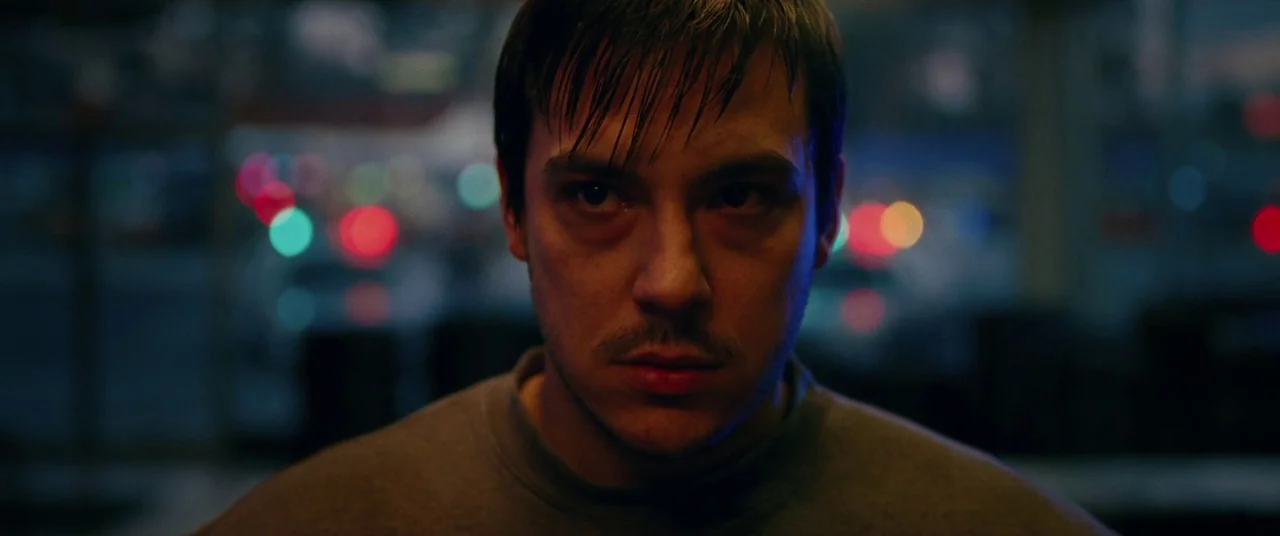 I, Tonya (2017) movie still: closeup — A man stands in a dark commercial space at night with colorful bokeh lights behind. He stares forward…
