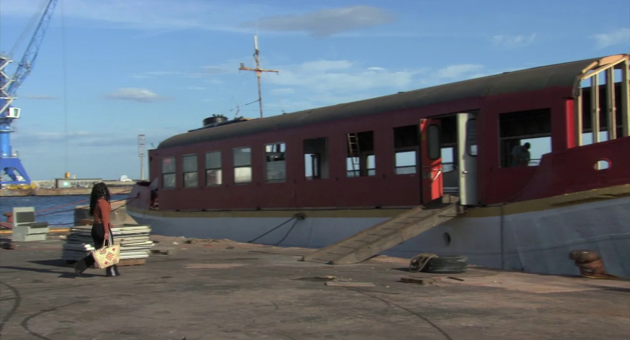 The Secret of the Grain (2007) movie still: wide — A woman walks along a weathered concrete pier near a large red boat vessel. A industrial crane…