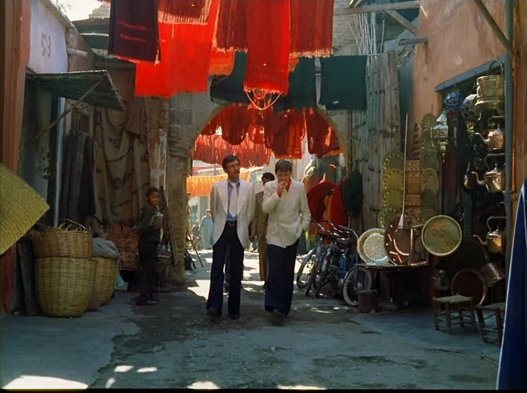 Fox and His Friends (1975) movie still: fullbody — A narrow market passage is filled with hanging red fabric and stacked copper goods. Two men walk…