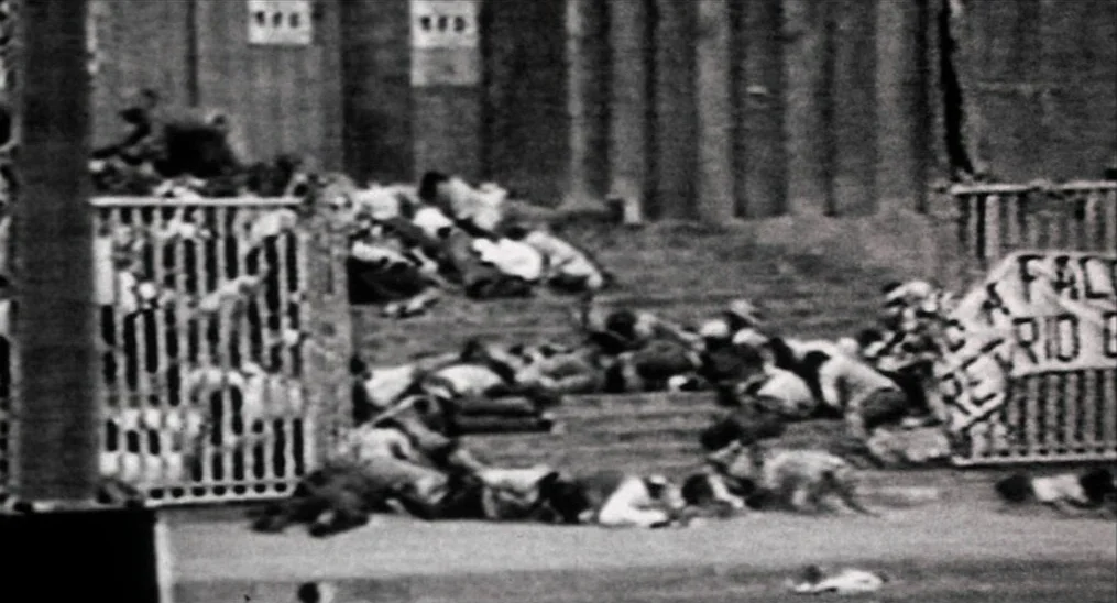Salvador (1986) movie still: wide — A crowded public space with multiple people lying prone on the stone ground. Metal fences and banners are…