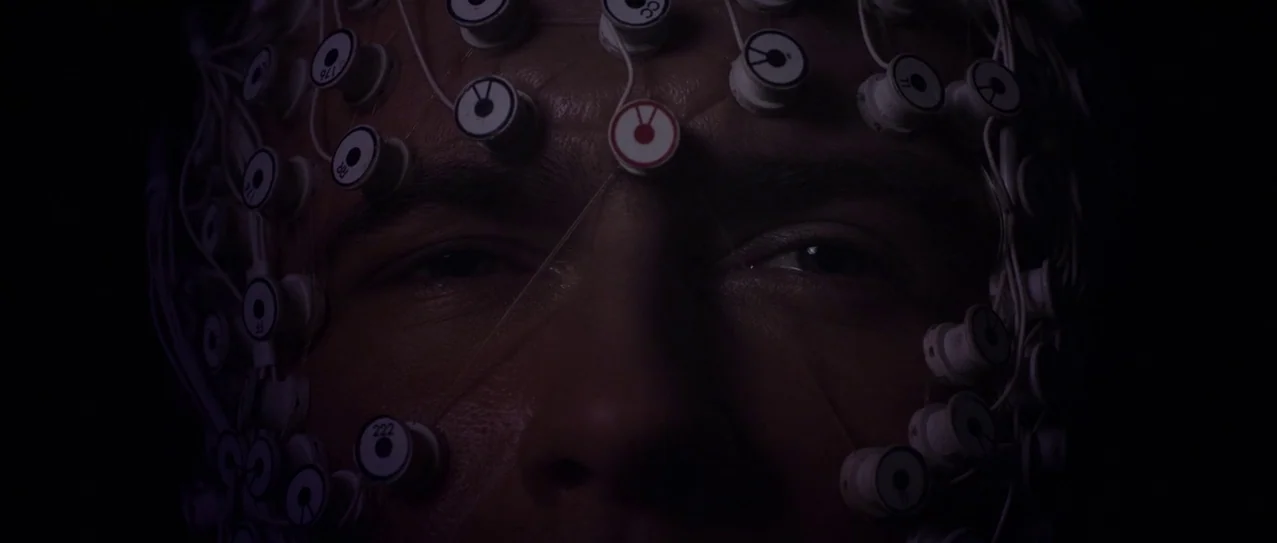 Vanishing Waves (2012) movie still: closeup — A dimly lit room focused on a man's face covered in medical sensors. The man stares ahead with his…