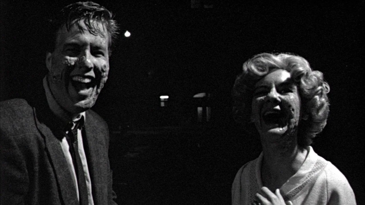 Poison movie still: medium — A dark, empty street at night is illuminated only by distant artificial lights. A man and woman stand in the frame,…