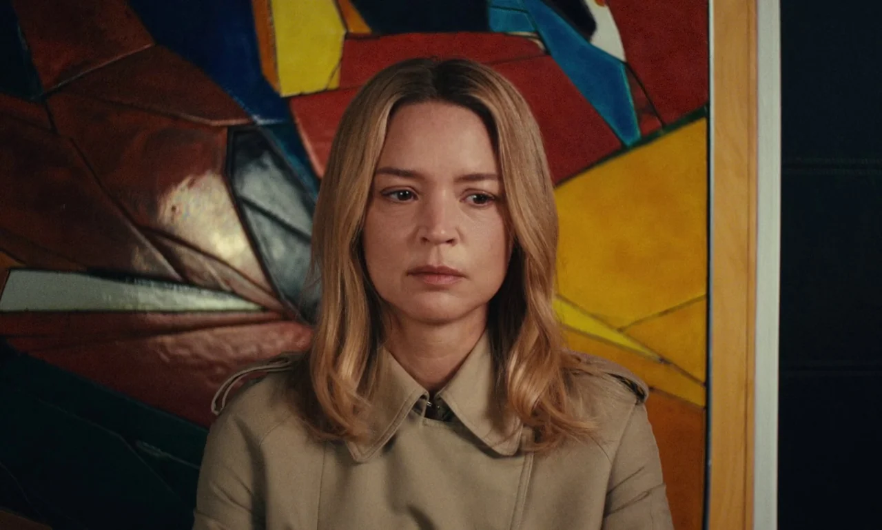 Just The Two Of Us (2023) movie still: closeup — A woman stands before a large, abstract stained glass panel with angular colorful shapes. She faces…