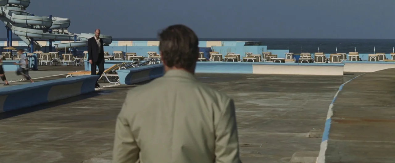 Syriana (2005) movie still: medium — An outdoor pool complex features a large blue water slide and many lounge chairs. Two men stand on the concrete…