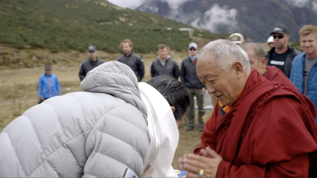 Drop Zone Everest (2023) movie still: medium — A high-altitude mountainous region with grassy slopes and cloudy peaks in the distance. A Buddhist…