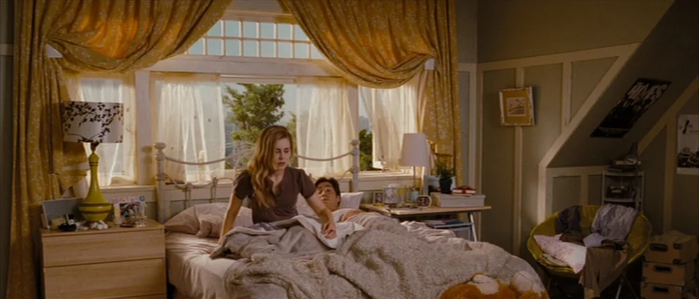 Drag Me to Hell (2009) movie still: medium — A well-lit bedroom features yellow curtains over a large window and wooden furniture. A woman sits up…