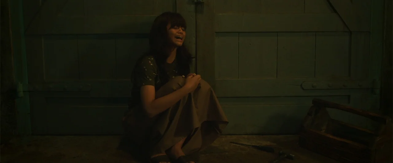 The Unholy (2022) movie still: medium — A dark indoor space with a large wooden door and concrete floor. A young woman crouches on the ground,…