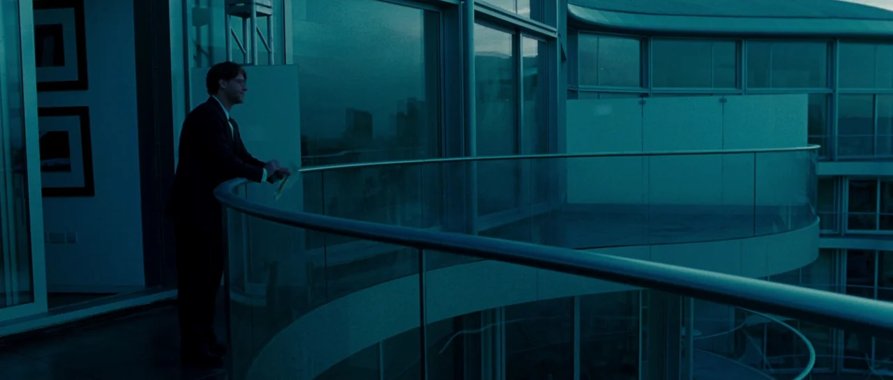 A Good Year (2006) movie still: fullbody — A modern curved glass balcony extends from a high-rise building at night. A man in a dark suit stands by…