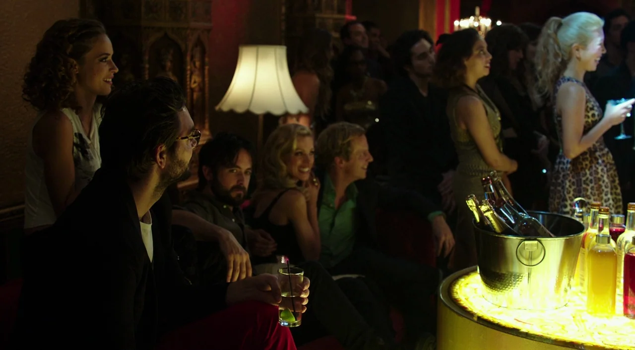 Urge (2016) movie still: medium — A dimly lit lounge area features a glowing circular bar with bottles of liquid. A group of people sit on red sofas…