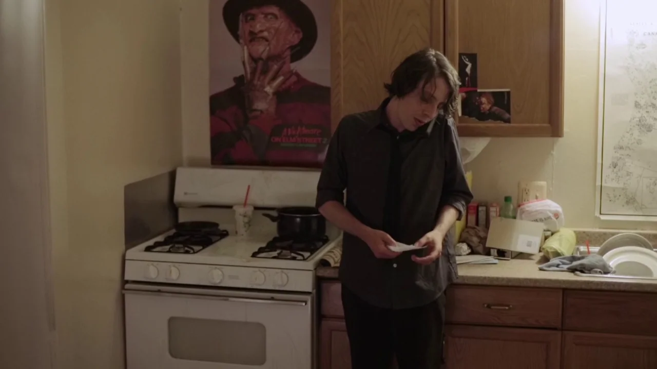 Buzzard (2014) movie still: medium — A modest home kitchen features wood-grain cabinets and a vintage white electric stove. A young man talks on a…