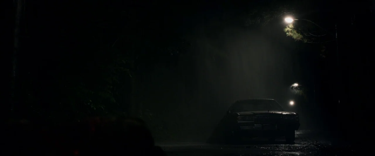 The Monster (2016) movie still: wide — A dark, wet road winds through a heavily wooded area at night. A car is parked underneath a single…