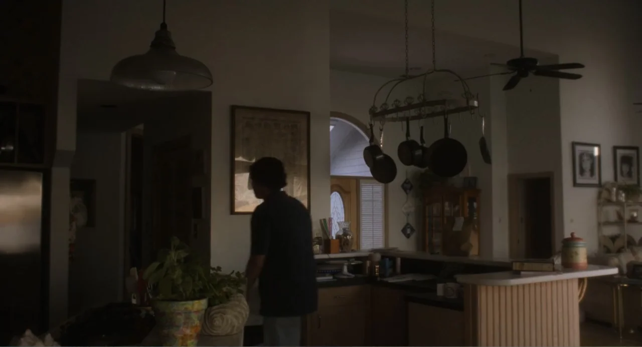 May December (2023) movie still: medium — A residential interior features a pot rack hanging above a kitchen island counter. A person wearing a dark…