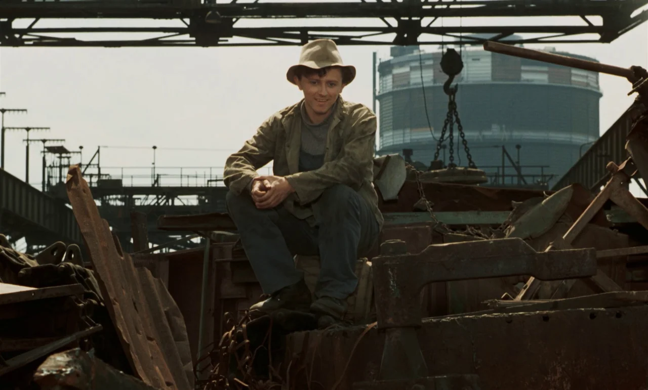 Larks On A String (1969) movie still: fullbody — An industrial junkyard features rusted metal debris and a large circular gas storage tank. A young…
