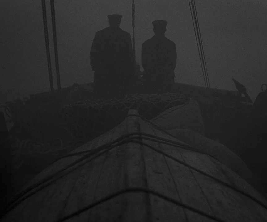 The Lighthouse (2019) movie still: wide — Two men in coats and peaked caps stand on a wooden boat deck. Heavy fog surrounds the vessel while the two…