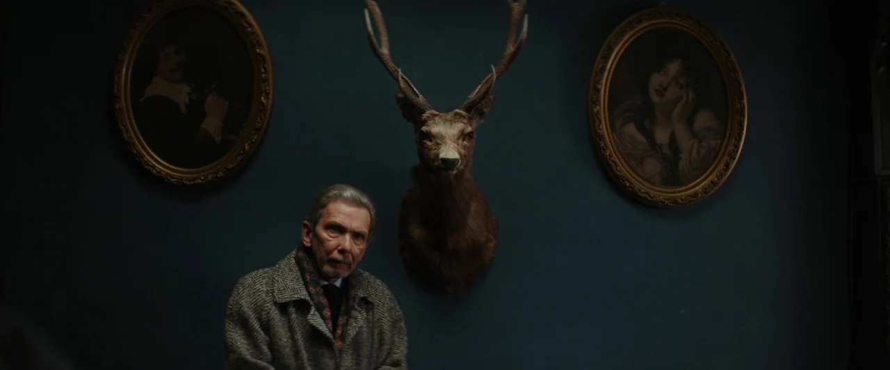Extra Ordinary (2019) movie still: medium — A dark teal wall displays two oval framed paintings flanking a mounted deer head. An elderly man in a…
