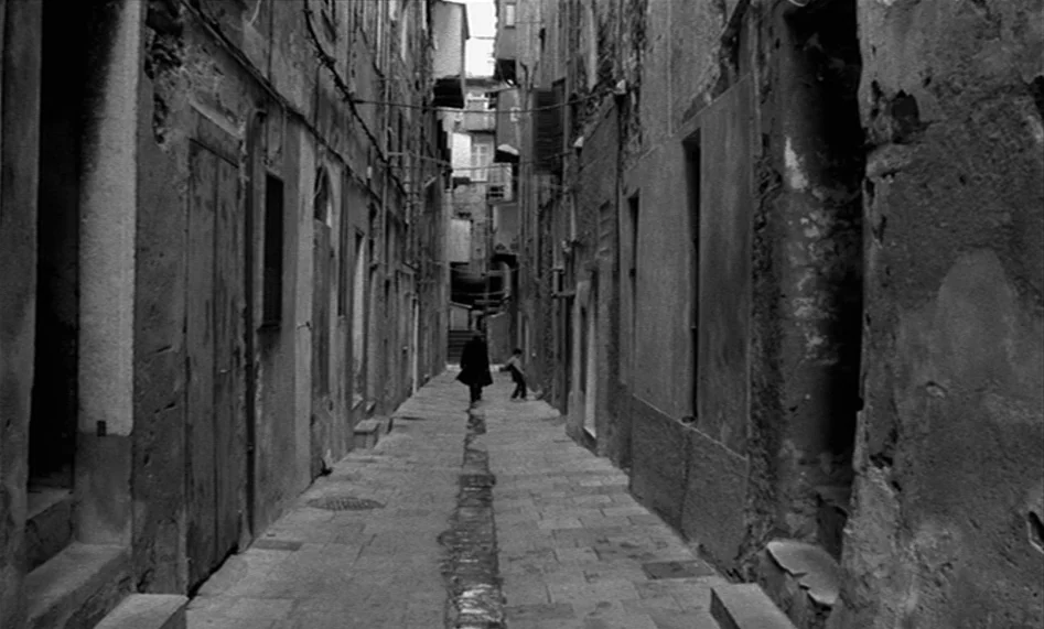 The Man From London (2007) movie still: wide — A narrow, stone-paved alleyway is flanked by tall, weathered buildings with peeling plaster walls. A…
