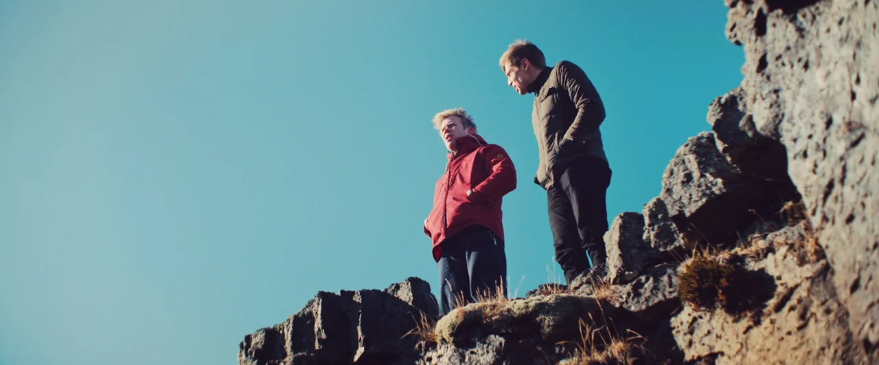 Rift (2017) movie still: fullbody — Two men stand atop a rocky, mossy outcrop against a clear, solid blue sky. They look outward while standing…