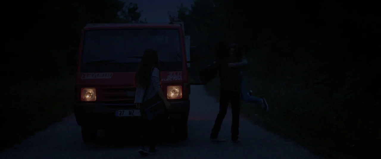 Mustang (2015) movie still: fullbody — A rural asphalt road at night is occupied by a stationary red commercial truck. Three figures stand near the…