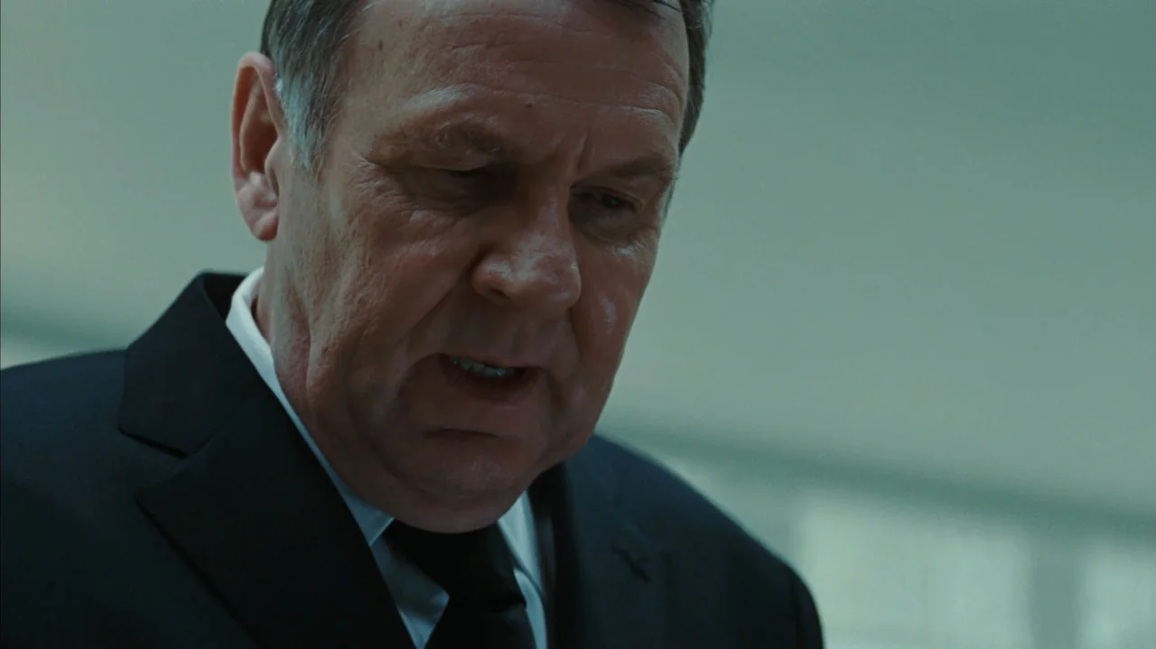 Duplicity (2009) movie still: closeup — A man in a suit stands in a sterile, dimly lit professional office environment. The subject looks downward…