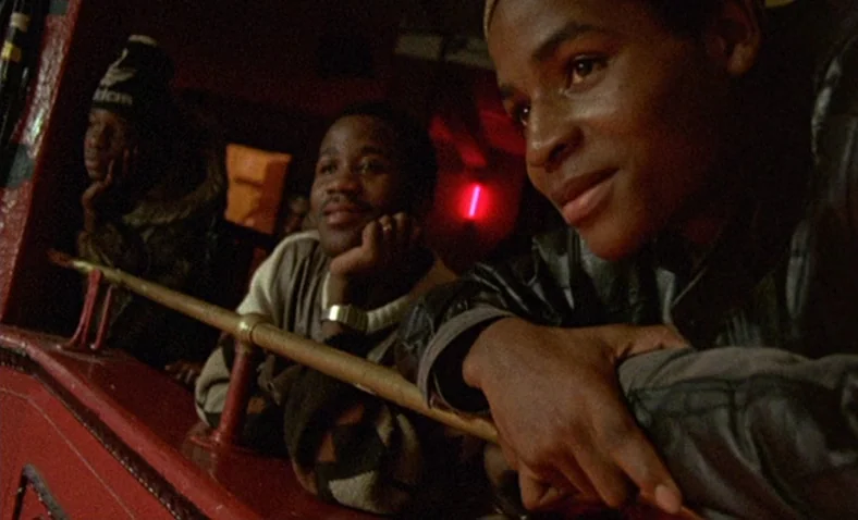 Paris Is Burning (1990) movie still: medium — A dimly lit theater balcony features red structural elements and a wooden railing. Three young men…
