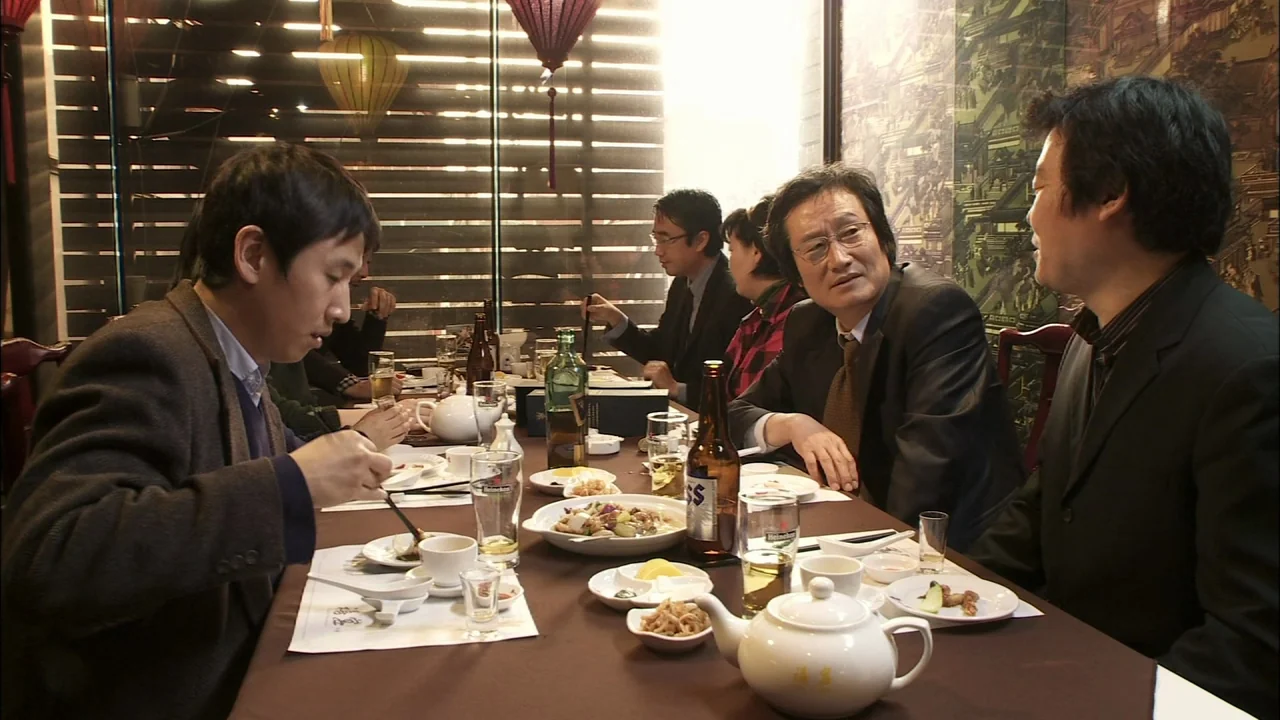 Oki’s Movie (2010) movie still: medium — A group of men sit at a long dining table inside a restaurant. They are sharing a meal with plates of food…