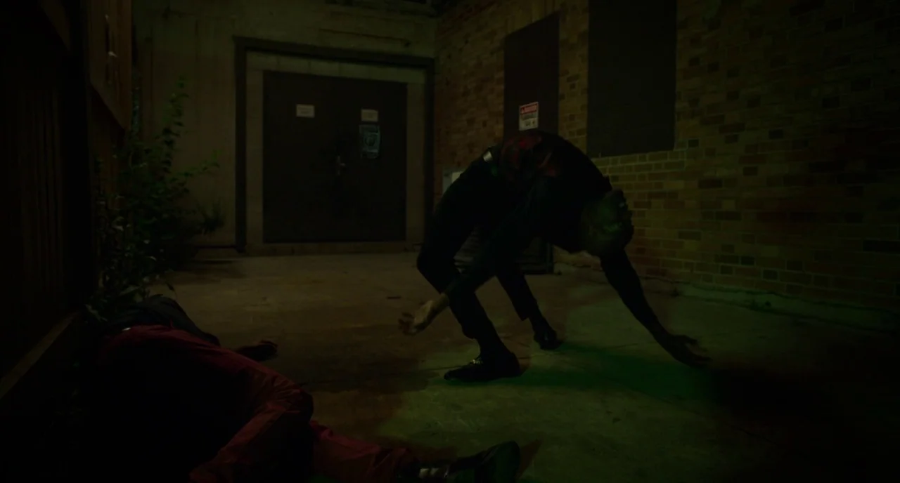 Rabid (2019) movie still: fullbody — A dark outdoor space beside a brick wall features a metal door and concrete. A standing figure arches their…