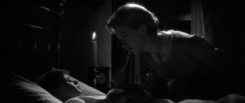 The Innocents (1961) movie still: medium — A dark, candlelit space with a bed frame and a clock visible in shadow. A woman leans over a child lying…