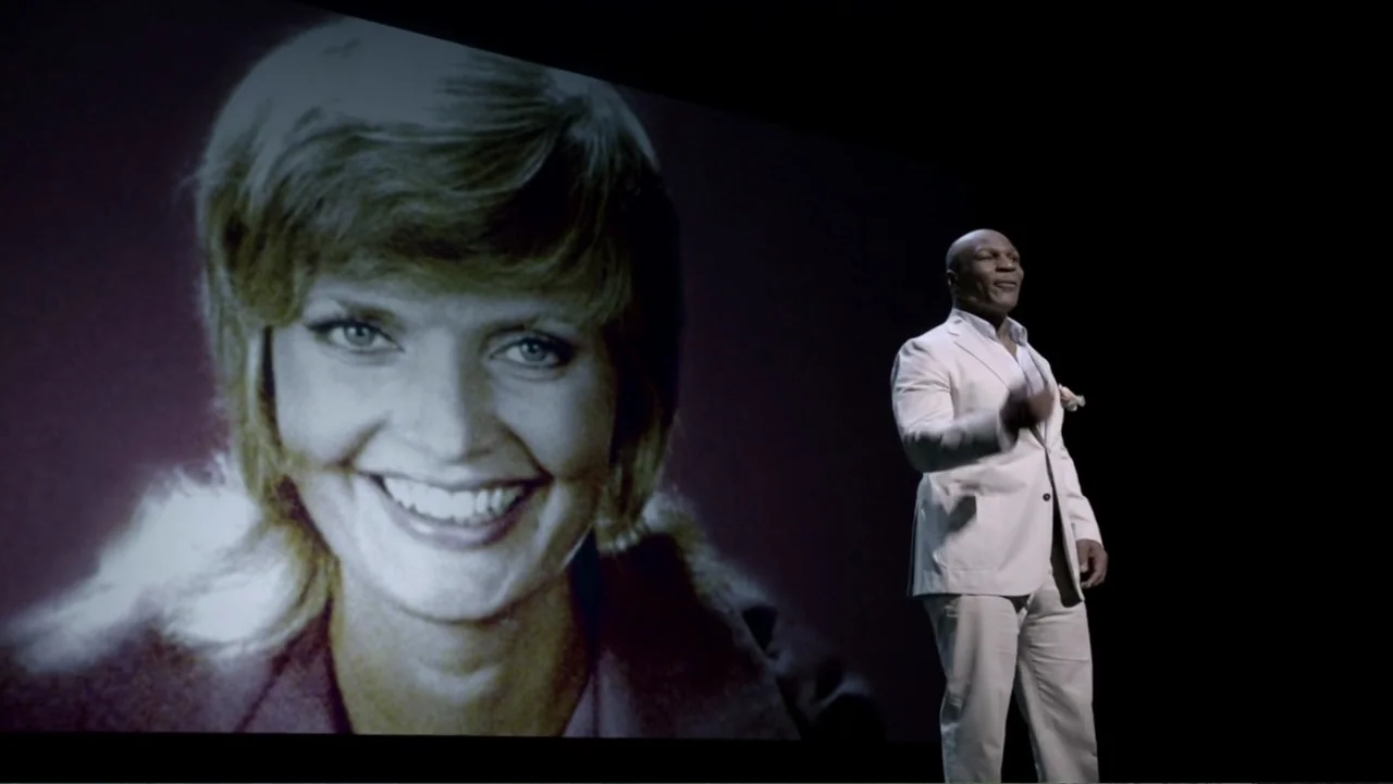 Mike Tyson: Undisputed Truth (2013) movie still: medium — A dark, empty stage features a large projection of a smiling woman behind a speaker. The…