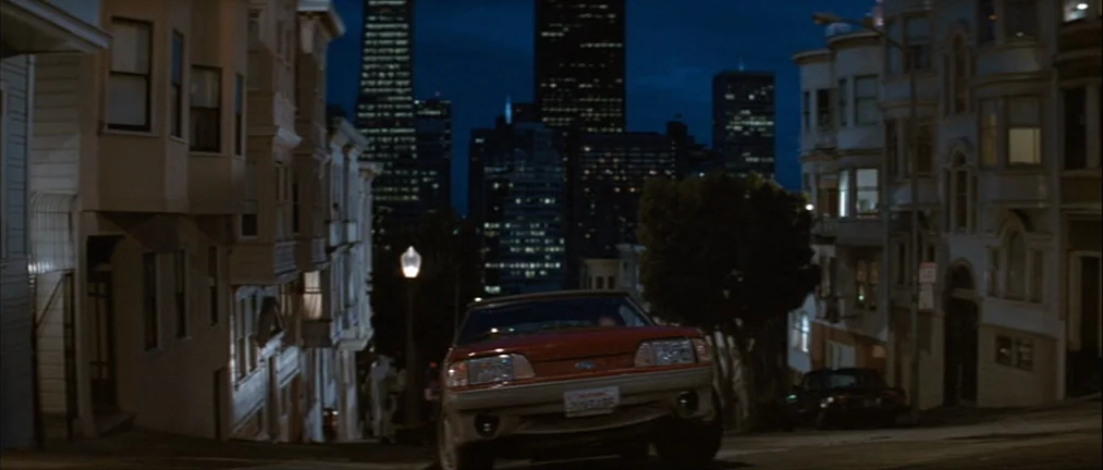 Basic Instinct (1992) movie still: wide — A steep residential street in San Francisco features Victorian houses on both sides at night. A red car…