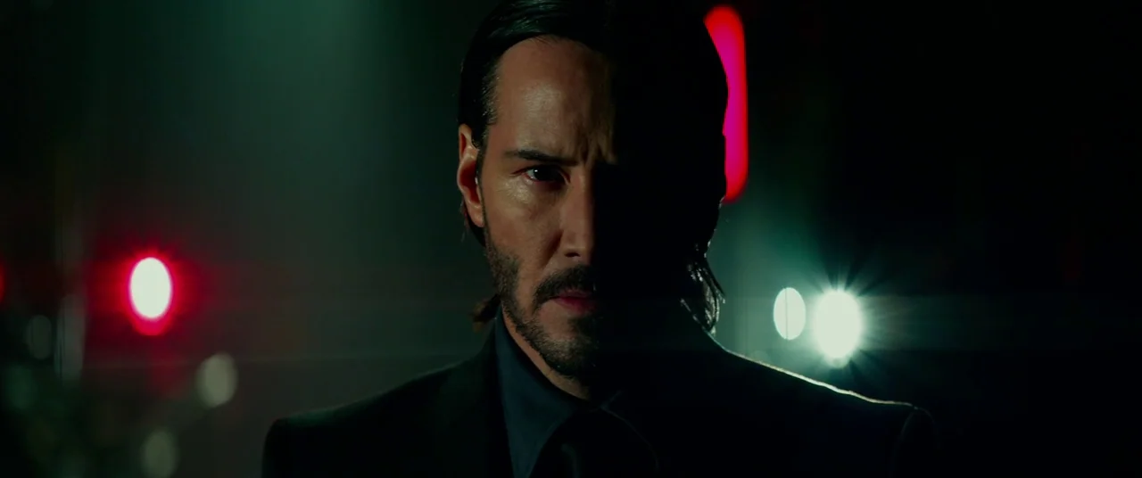 John Wick: Chapter 2 (2017) movie still: closeup — A dark city street at night illuminated by red and white blurred neon lights. A man in a dark…