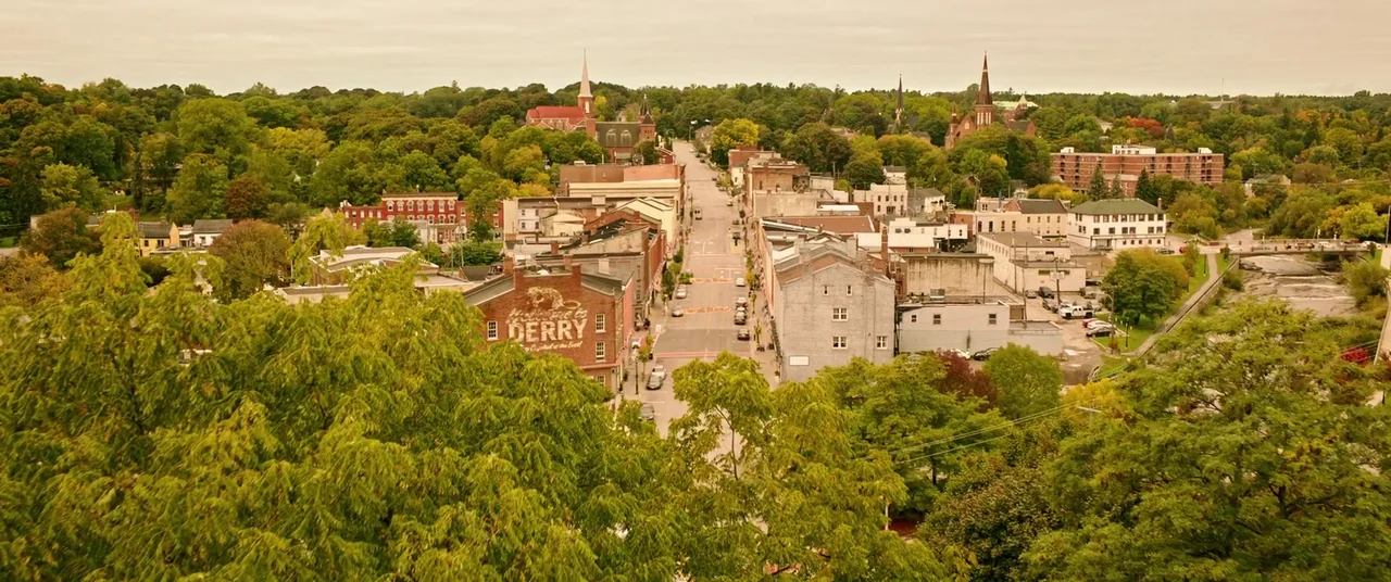 It Chapter Two (2019) movie still: establishing — An aerial view of a small town features a central road flanked by brick buildings. Lush green…